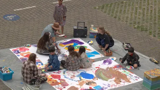 Kinder und Jugendliche sitzen auf einer Plane mit großem, weißen Papier. Drum herum Kunst-Utensilien mit denen sie dieses Papier bunt bemalen. Sie tragen Malerkittel und sind auf einem Parkplatz.