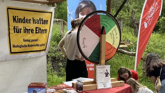 Se puede ver un puesto de información con techo y muchos folletos sobre la mesa. Junto a él hay una caja de donaciones y una ruleta de premios de Terre des Hommes . Unos niños juegan en el suelo, cerca de allí.