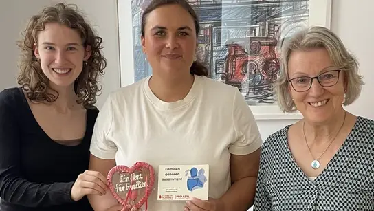 Tres mujeres están de pie una junto a la otra frente a un cuadro en una oficina. Dos de ellas son miembros del grupo Terre des Hommes de Heidelberg-Mannheim. Entre ellas se encuentra la política Isabelle Cademartori. Sostiene en la mano un corazón de pan de jengibre con la inscripción "Un corazón para las familias" y un folleto de la campaña.