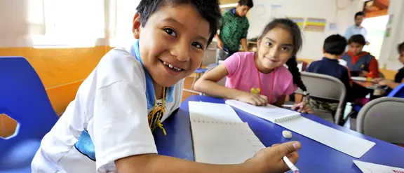 Ein Junge und ein Mädchen arbeiten an einem blauen Tisch, im Hintergrund sind weitere Kinder