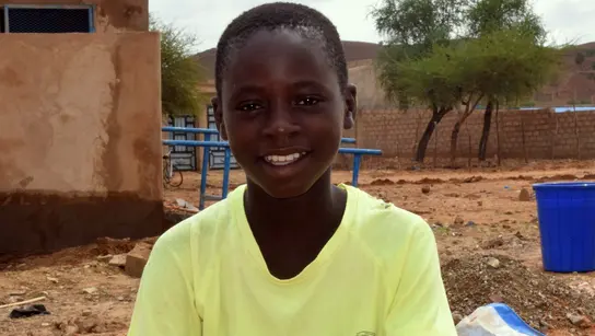 Lächelnder Junge sitzt vor einem Gebäude in einer ländlichen Gegend von Burkina Faso, umgeben von einfacher Architektur und weiter Landschaft.
