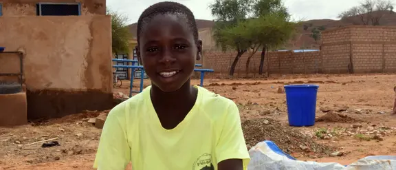 El niño sonriente se sienta frente a un edificio en una zona rural de Burkina Faso, rodeado de arquitectura simple y un paisaje adicional.
