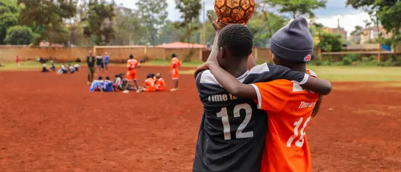 Zwei Kinder mit einem Fußball vor im Vordergrund, im Hintergrund weitere Kinder mit Fußballkleidung