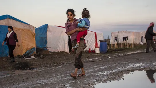 Ein Mädchen trägt ein kleines Kind. Im Hintergrund sind Zelte und weitere Menschen zu sehen