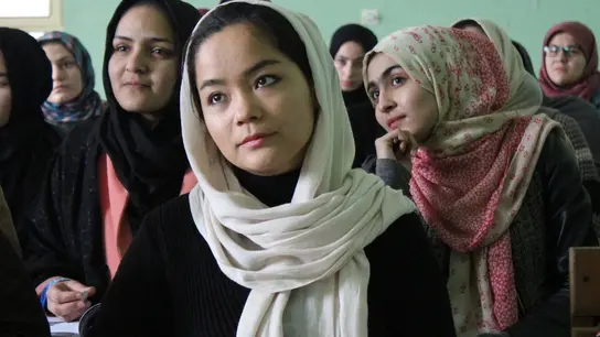 Junge Frauen sitzen in einem Klassenzimmer, tragen Kopftücher und halten Bücher. Sie sind aufmerksam und konzentriert, während sie dem Unterricht folgen.