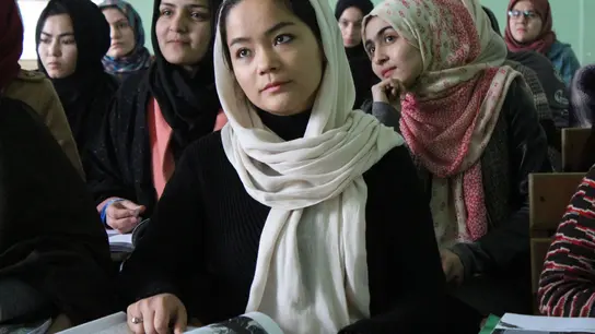 Junge Frauen sitzen in einem Klassenzimmer, tragen Kopftücher und halten Bücher. Sie sind aufmerksam und konzentriert, während sie dem Unterricht folgen.