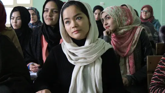 Junge Frauen sitzen in einem Klassenzimmer, tragen Kopftücher und halten Bücher. Sie sind aufmerksam und konzentriert, während sie dem Unterricht folgen.