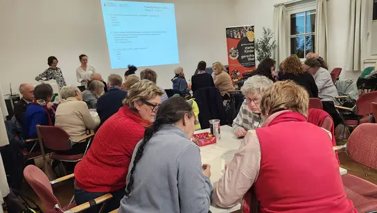 Personen sitzen an Tischen und beraten sich. Im Hintergrund ist eine Präsentationsfolie an die Wand geworfen mit Quizfragen