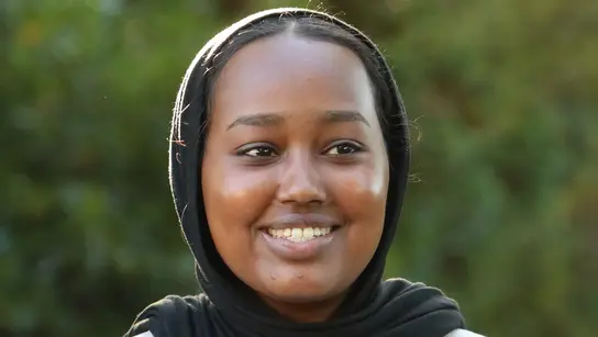 Ein Mädchen aus Somalia namens Sainab lächelt und spricht. Sie trägt ein schwarzes Kopftuch und ein weißes T-Shirt.