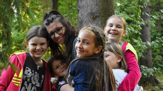 Five girls and a woman stand in a forest, smiling and hugging each other 