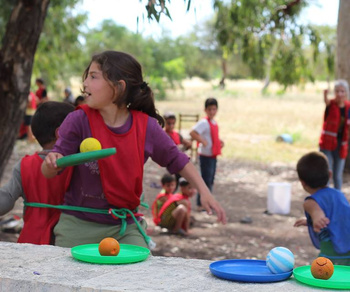 Ein Mädchen hat eine Frisbee Scheibe und einen Ball in der Hand, Kinder spielen im Hintergrund.