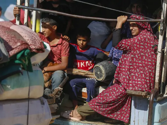 Kinder und eine Frau sitzen eng beieinander auf der Ladefläche eines beladenen Fahrzeugs mit Decken und Gepäck – Flüchtende aus Gaza in einer humanitären Notsituation.