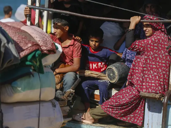 Kinder und eine Frau sitzen eng beieinander auf der Ladefläche eines beladenen Fahrzeugs mit Decken und Gepäck – Flüchtende aus Gaza in einer humanitären Notsituation.