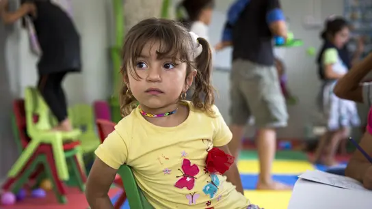 Ein junges Mädchen sitzt vor einem Tisch, an dem gemalt wird, im Hintergrund spielen Kinder