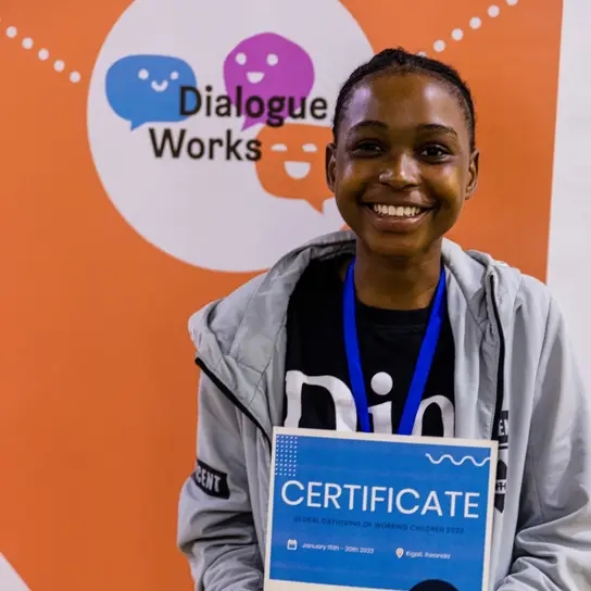 Ein Mädchen zeigt lächelnd ihr Zertifikat auf einer Konferenz. Im Hintergrund ein Schild auf dem "Dialogue Works" steht.