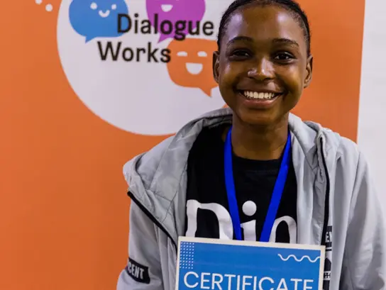 Ein Mädchen zeigt lächelnd ihr Zertifikat auf einer Konferenz. Im Hintergrund ein Schild auf dem "Dialogue Works" steht.