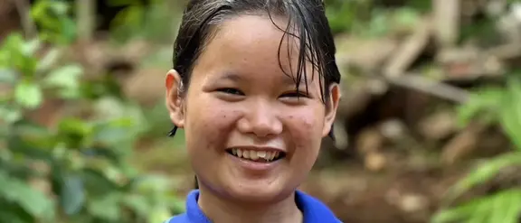 Ein junges Mädchen aus Myanmar namens Melada steht vor einem grünen Wald und lächelt für das Portraitbild. Sie trägt ein blaues Polo-T-Shirt.