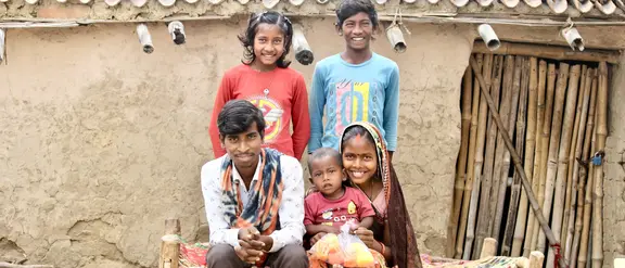 Eine indische Familie sitzt vor einem Lehmhaus, wobei die Eltern mit ihrem Kleinkind auf einer Bank sitzen und die älteren Kinder dahinter stehen.