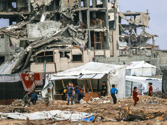 Eine Trümmerlandschaft in Gaza. In der Mitte des Bildes sind spielende Kinder aus der Ferne zu sehen.