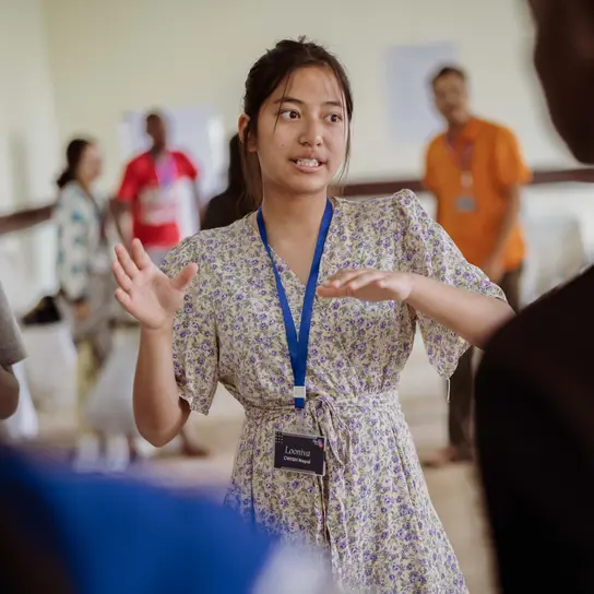Jugendliche aus Nepal spricht und gestikuliert dabei mit den Händen. Sie trägt ein Kleid und hat ein Schlüsselband um den Hals.