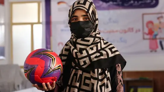Mujer joven en un pañuelo y máscara estampado contiene un fútbol colorido en una habitación con una pancarta en el fondo.