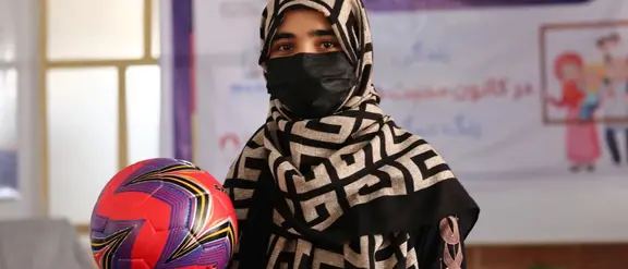 Mujer joven en un pañuelo y máscara estampado contiene un fútbol colorido en una habitación con una pancarta en el fondo.