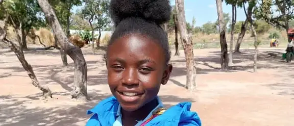 Ein junges Mädchen aus der Demokratischen Republik Kongo namens Manda posiert lächelnd für das Portrait. Sie trägt eine hellblaue Jacke und steht vor dem Hintergrund einer Steppenlandschaft.