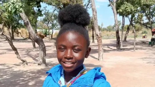 Ein junges Mädchen aus der Demokratischen Republik Kongo namens Manda posiert lächelnd für das Portrait. Sie trägt eine hellblaue Jacke und steht vor dem Hintergrund einer Steppenlandschaft.
