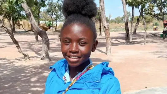 Ein junges Mädchen aus der Demokratischen Republik Kongo namens Manda posiert lächelnd für das Portrait. Sie trägt eine hellblaue Jacke und steht vor dem Hintergrund einer Steppenlandschaft.