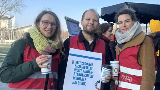 Drei Terre des Hommes Mitarbeitende mit einem Schild im Vordergrund, im Hintergrund weitere Personen