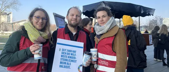 Drei Terre des Hommes Mitarbeitende mit einem Schild im Vordergrund, im Hintergrund weitere Personen