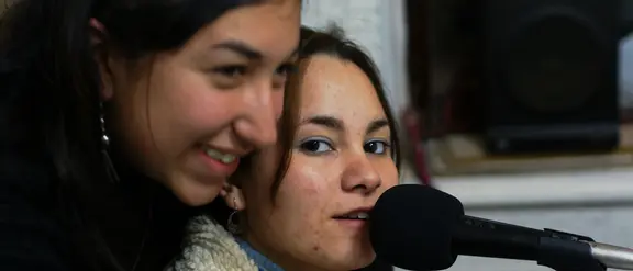 Zwei junge Frauen in einem Tonstudio, eine spricht in ein Mikrofon, die andere lächelt daneben.