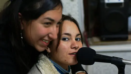 Dos mujeres jóvenes en un estudio de grabación, uno habla en un micrófono, el otro sonríe al lado.