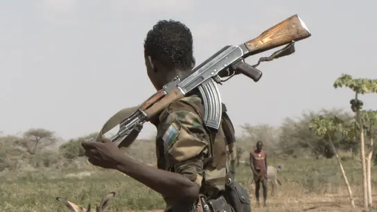 Ein junger Mensch mit einer Waffe, dahinter ein Esel