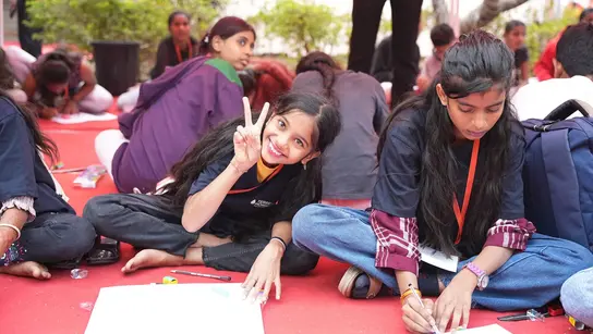 Zwei indische Mädchen sitzen mit Schulbüchern auf dem Boden. Eines schaut und lacht den Betrachter an.