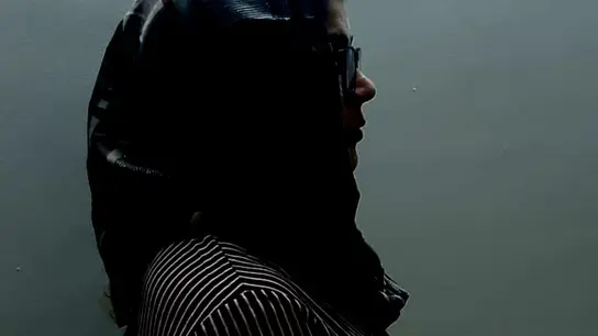 Ein Profilbild einer Frau mit Kopftuch und Brille vor einem schlichten Hintergrund.