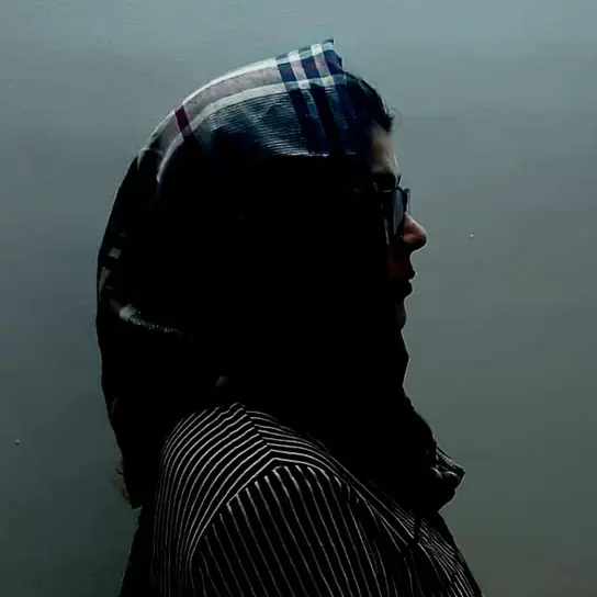 Ein Profilbild einer Frau mit Kopftuch und Brille vor einem schlichten Hintergrund.