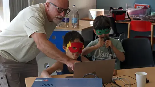 Zwei Jungen tragen selbst gebastelte Superhelden-Masken. Der eine steht an einem Laptop, der andere rappt in ein Mikrofon. Ein älterer Herr steht daneben und zeigt ihnen etwas auf dem Laptop.