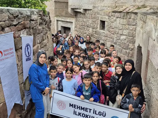 Zwischen alten Stadtmauern steht eine große Gruppe von Kindern in der Türkei, die eine Wanderung machen. Betreuerinnen stehen daneben und sie halten Plakate und Banner in der Hand.