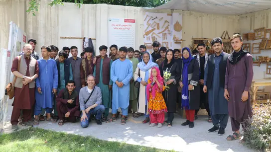 Eine Gruppe von Menschen in traditioneller Kleidung steht vor einer Wand mit Werkzeugen und Schildern. Die Szene findet im Freien statt, umgeben von grüner Vegetation.