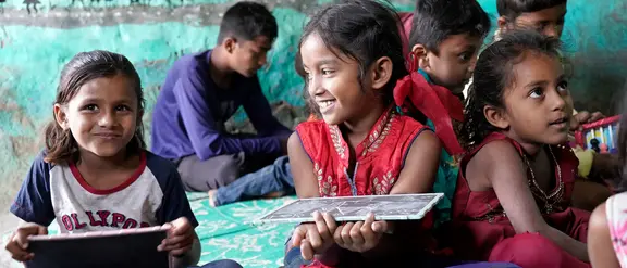Lachende Kinder in einem Klassenzimmer in Indien sitzen auf dem Boden und halten Tafeln. Die Szene zeigt eine lebendige und kreative Lernumgebung, die die Freude am Lernen und die kulturelle Vielfalt in Indien betont.