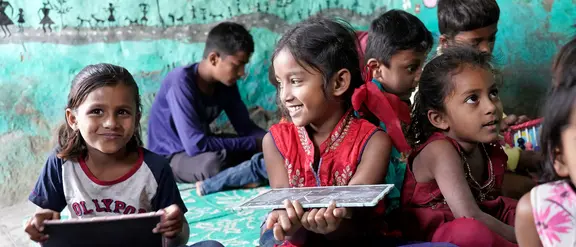 Laughing children in a classroom in India sit on the floor and hold boards. The scene shows a lively and creative learning environment that emphasizes the joy of learning and cultural diversity in India.