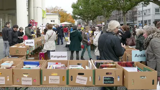 Personen gehen entlang an einer Reihe an Kisten mit Büchern