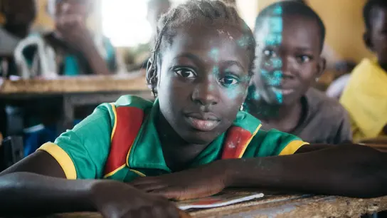 Una niña atenta se sienta en un salón de clases en Burkina Faso. La escena muestra la importancia de la educación y la esperanza de un futuro mejor a través del aprendizaje.