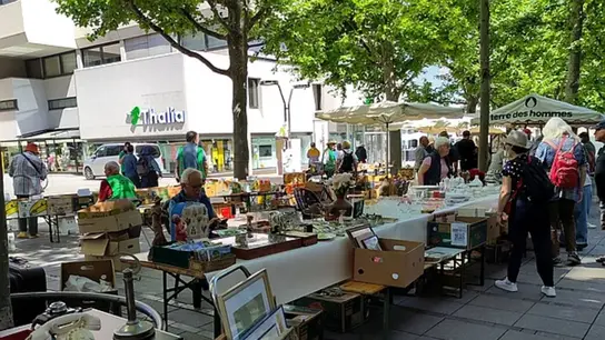 Bild zeigt, Flohmarktgegenstände auf und unter Tischen. Im Hintergrund schauen sich Passanten die Gegenstände an.