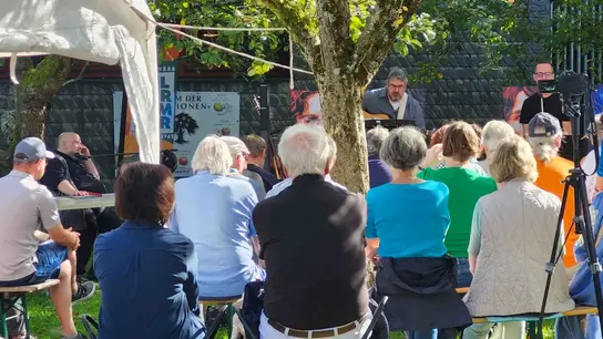 Personen sitzen auf Bierbänken an einem sommerlichen Nachmittag und hören einem Konzert zu.