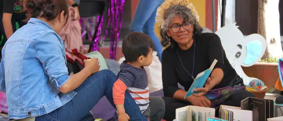 Eine Frau zeigt einem kleinen jungen ein Buch, links im Bild sind zwei weitere Frauen von der Kamera abgewandt