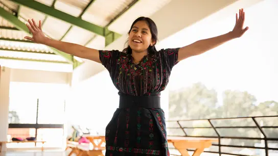 Ein Mädchen aus Guatemala steht auf einer Terasse und streckt beide Arme aus als würde sie fliegen. Sie lacht.