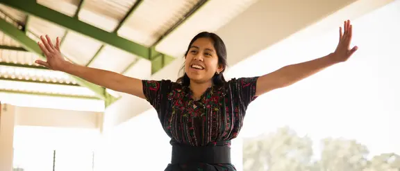 Ein Mädchen aus Guatemala steht auf einer Terasse und streckt beide Arme aus als würde sie fliegen. Sie lacht.