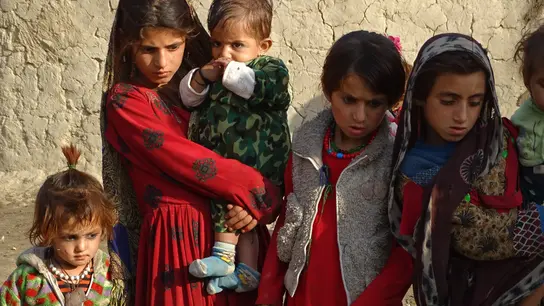 Eine Gruppe von Kindern in bunter, traditioneller Kleidung steht vor einer lehmigen Wand.